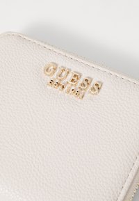 Grijze textuurportemonnee met gouden "GUESS" logo en "EST 1981" detail, voorzien van een ritsluiting en zichtbare stiksels op de randen.