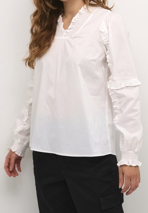 Blusa bianca a maniche lunghe con bordi arricciati su colletto, maniche e polsini, indossata con pantaloni neri.