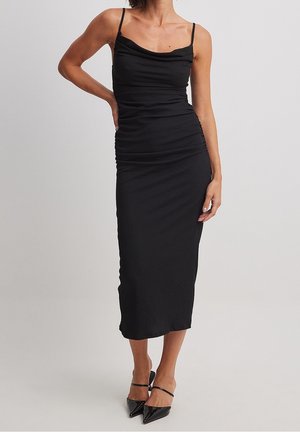 Schwarzes Midi-Kleid mit dünnen Trägern und einem schalenförmigen Ausschnitt. Der Stoff ist strukturiert und schmiegt sich mit gerafften Details an den Körper. Kombiniert mit spitzen schwarzen High Heels.