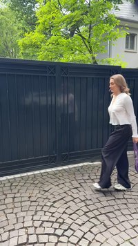 Una persona in camicia bianca con bottoni e pantaloni neri a gamba larga passa accanto a un cancello in metallo scuro. Il sentiero di ciottoli presenta un motivo curvo.