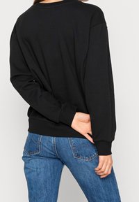 Personne portant un pull noir à manches longues et un jean bleu, avec une main posée derrière le dos.