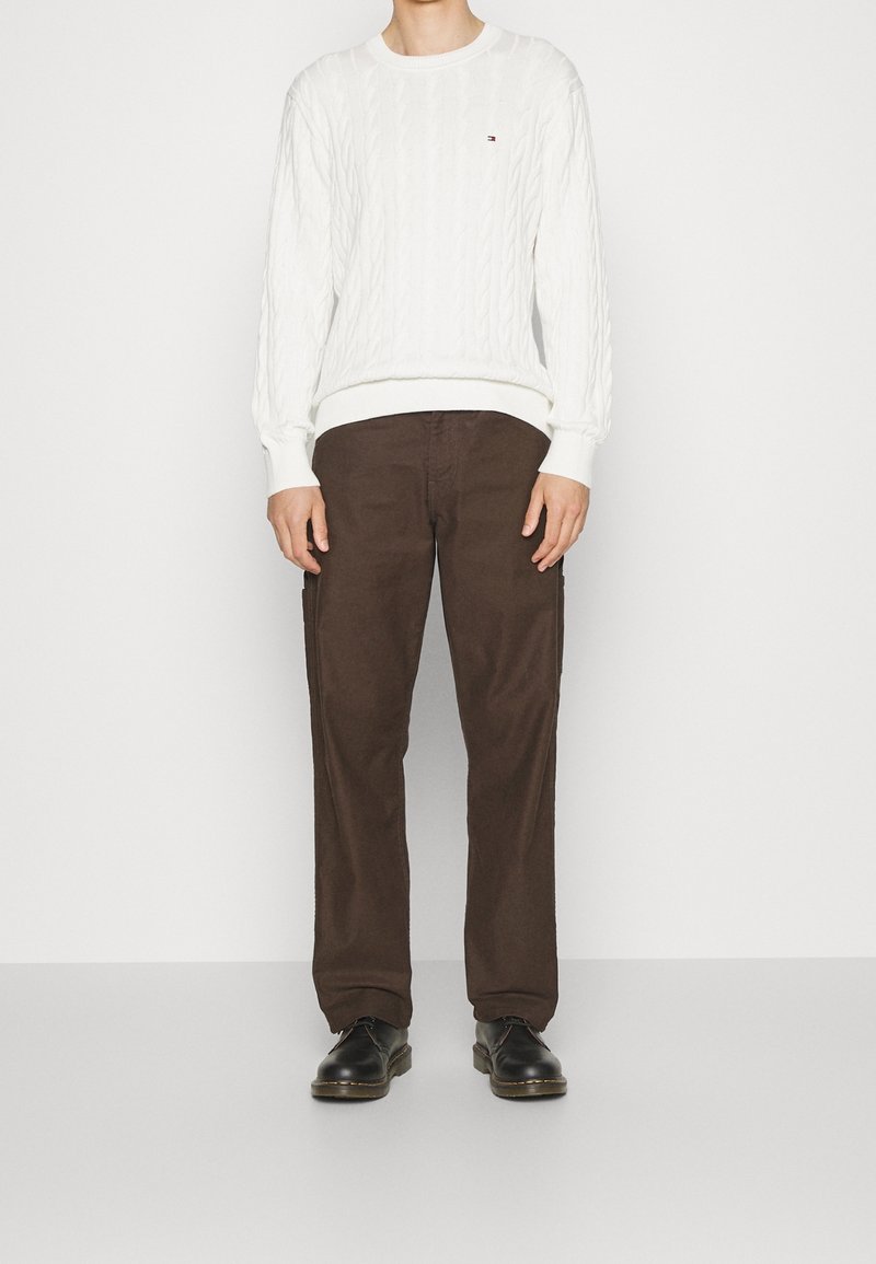 Personne portant un pull en maille torsadée blanc, un pantalon marron et des chaussures noires, debout devant un fond blanc uni.