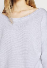 Maglione lavorato a maglia di colore grigio chiaro, di grandi dimensioni, con un ampio scollo a costine e tessuto strutturato. Presenta una vestibilità rilassata, con spalle leggermente cadenti.