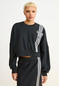 Melna īsa sporta jaka ar apaļu kakla izgriezumu, baltu Adidas logo uzplecu un trim baltām svītrām pa kreiso piedurkni. Mīksta tekstūra.
