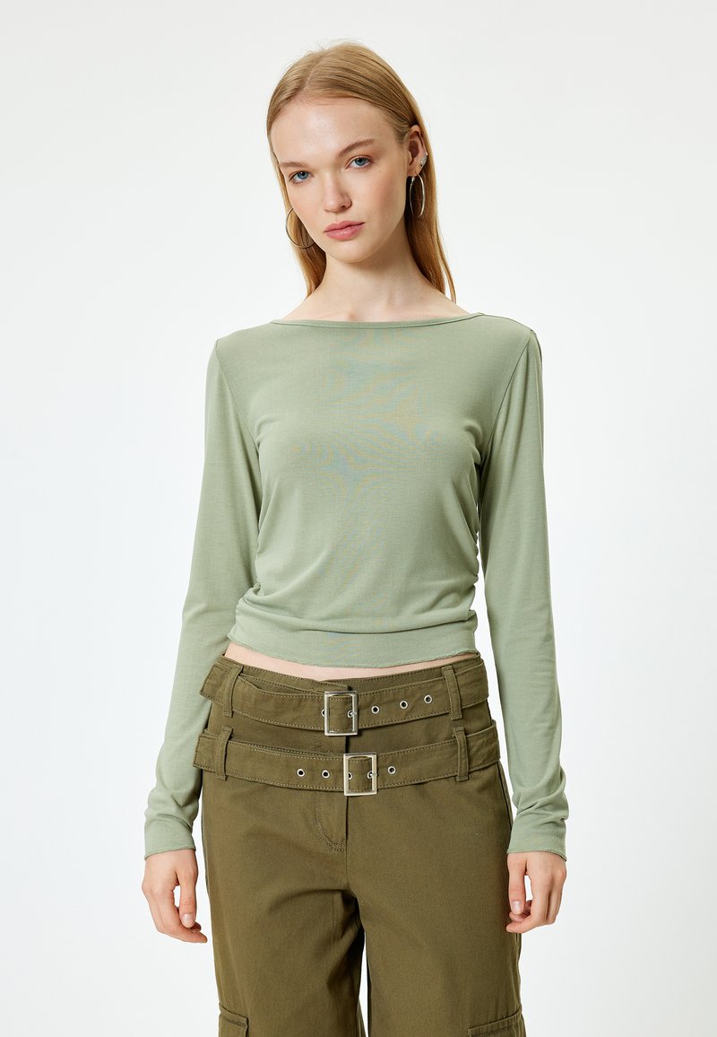 Hellgrünes Langarmshirt mit Bootsausschnitt, kombiniert mit olivgrünen Cargo-Hosen, die über doppelte Gürtelschnallen und große Gürtelschlaufen verfügen.