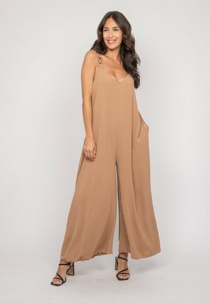 Femme aux longs cheveux foncés portant une combinaison ample sans manches de couleur beige avec des jambes larges et des talons noirs à brides, debout devant un fond uni.