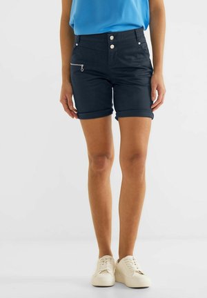 Vrouw die marineblauwe shorts met omgeslagen zoom en een voorvak met rits draagt, een blauw shirt en witte sneakers, staand tegen een effen achtergrond.