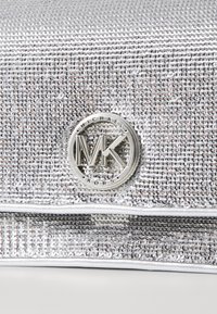 Sidabrinė blizgučiais dekoruota rankinė, turinti apvalų metalinį logotipą su "MK" centre ir "MICHAEL KORS" aplink jį. Lygi tekstūra.