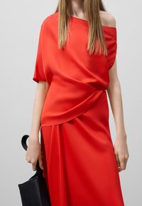 Abito di raso rosso con scollatura asimmetrica e design drappeggiato. Abbinato a una borsa nera, che mette in mostra una texture liscia e una silhouette fluida.