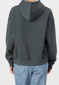 Sudadera con capucha en mezcla de algodón verde oscuro con un corte relajado. Presenta puños y dobladillo de canalé. Llevada con pantalones de patrón de rosa en azul claro.