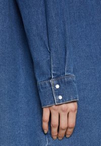 Denim shirt sleeve with a fitted design, blue color, and pearl snap buttons. Fabric shows a smooth texture and slight fading.