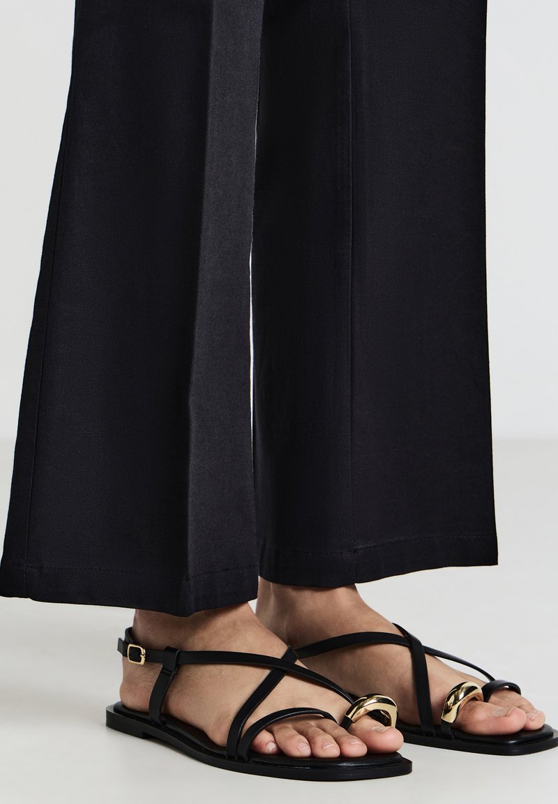 Pieds chaussés de sandales plates noires avec des détails en métal doré torsadé et des brides aux chevilles, associés à un pantalon noir à jambes larges.