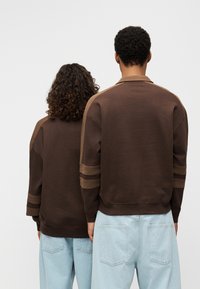 Καφέ φούτερ με χαλαρή εφαρμογή, διαθέτει ριμπ γιακά, ριγέ διακοσμητικά σε ανοιχτότερη απόχρωση και αντίθετο ανοιχτό μπλε τζιν στη μέση.