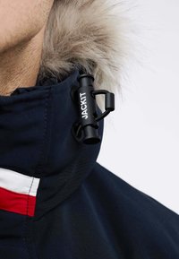Col de veste marine avec bordure en fausse fourrure, équipé d'un bouton de réglage noir marqué "JACK1T" et de bandes d'accent rouge et blanc.