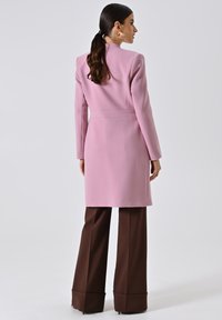 Cappotto rosa su misura con colletto alto e maniche lunghe, presenta un design senza cuciture e una vita aderente. Abbinato a pantaloni marroni larghi.