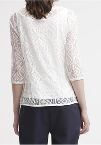 Bílý krajkový top s rukávy do tří čtvrtin, který má geometrický vzor, texturovanou tkaninu a poloprůhledný design na spodním lemu.