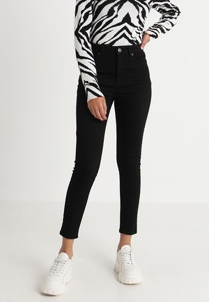 Kvinde iført sorte, højtaljede skinny jeans, zebra-print langærmet top og hvide chunky sneakers på ensfarvet baggrund.