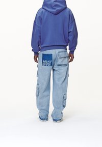 Felpa viola con cappuccio, dal fit rilassato e polsini a costine, abbinata a jeans cargo azzurri chiari con applicazioni grafiche e la scritta "NON È MAI TROPPO TARDI".
