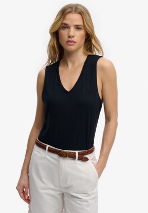 Vrouw die een zwarte, mouwloze top met V-hals draagt, ingestopt in een witte broek met een bruine gevlochten riem, staand met één hand in haar zak.