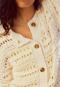 Cardigan en crochet blanc avec un motif en tricot ajouré, cinq grands boutons ronds et des accents texturés, superposé sur un haut jaune clair.