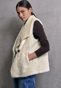 Gilet in pelliccia sintetica color crema con colletto, chiusura frontale a bottoni e tasche laterali. Indossato sopra una camicia scura a maniche lunghe e abbinato a jeans blu.