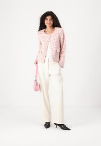 Rosa og hvit rutete blazer med gullknapper, kombinert med hvite stripete bukser. Holder en liten rosa håndveske; svarte spisse sko.