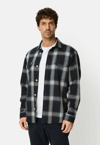 Koszula flanelowa w czarną i granatową kratę, z guzikami, dwoma kieszeniami na piersi oraz luźnym krojem. Noszona na białą koszulkę, z długimi rękawami.