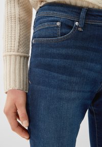 Mörkblå denimjeans har en mjuk textur, femfickorsdesign och synliga sömnadsdetaljer. En hand vilar på sidan.