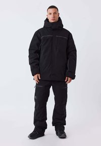 Zwart gewatteerd jack met capuchon, voorzien van contrasterende witte stiksels, met ritsen afsluitbare zakken en bijpassende zwarte broek, aangevuld met zwart schoeisel.