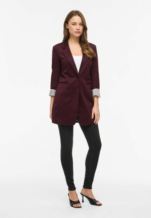 Junge Frau steht, trägt einen langen bordeauxroten Blazer mit aufgekrempelten Ärmeln, ein weißes Oberteil, schwarze Skinny-Jeans und schwarze Sandalen mit Absatz.