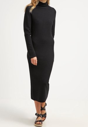 Femme portant une robe midi noire à manches longues côtelées, associée à des sandales à plateforme noires avec brides à la cheville.