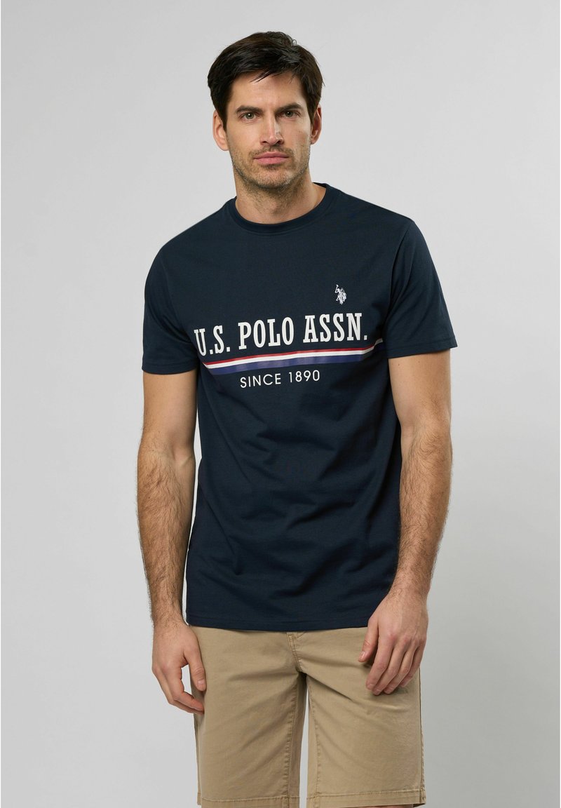 Majica od pamuka tamnoplave boje s kratkim rukavima, s bijelim logotipom i natpisom "U.S. POLO ASSN." te crveno-bijelim horizontalnim prugama.