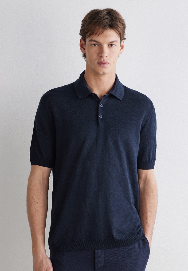 Junger Mann steht an einer weißen Wand, trägt ein dunkelblaues kurzärmeliges Poloshirt mit Kragen und Knöpfen, neutraler Gesichtsausdruck, Hände an den Seiten.