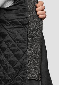 La fodera trapuntata nera mostra un tessuto grigio texturizzato al bordo, con una cerniera liscia con ferramenta metallica e un design aderente.