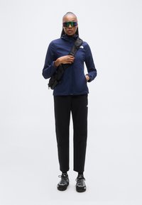 Μπλε τζάκετ με φερμουάρ και ψηλό γιακά, μαύρο παντελόνι και μαύρα παπούτσια. Τσάντα περασμένη στο σώμα, μεγάλα ανακλαστικά γυαλιά ηλίου ως λεπτομέρειες.