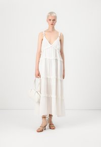 Hvit maksikjole med lagdelte volanger, V-hals og tynne stropper. Kombinert med en liten hvit håndveske og beigemiddels sandaler.