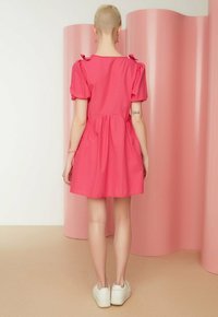 Roze korte jurk met pofmouwen, gathered taille en strikaccenten op de schouders. G gedragen met witte sneakers tegen een roze achtergrond.