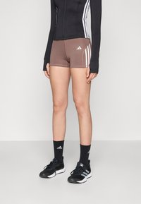 Brune idrettsshorts med tre hvite striper på sidene, kombinert med en svart langermet zip-up topp og svart fottøy med grå detaljer.