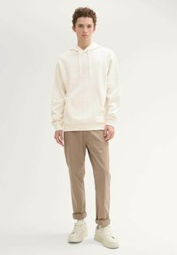 Kremowa bluza z kapturem z przednią kieszenią, ściągaczami na rękawach i sznurkiem; zestawiona z beżowymi spodniami i jasnymi sneakersami. Model stoi na neutralnym tle.