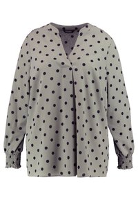Blouse verte à pois avec un col en V, texture douce, manches longues et poignets froncés. Elle possède une fermeture à boutons à l'avant.