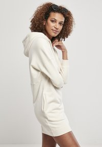 Rochie-sudor din material moale de culoare crem deschis, cu mâneci lungi, buzunare laterale și croială confortabilă, având o glugă cu șnur și manșete texturate.