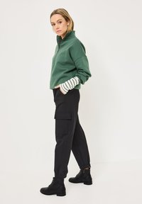 Femme se tenant de côté, portant un sweat à capuche vert zippé, une chemise à rayures à manches longues en dessous, un pantalon cargo noir et des bottes de cheville noires.