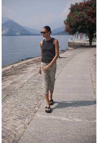 Haut sans manches noir, jupe midi à imprimé animal beige et sandales à plateforme noires. Femme marchant le long d'un chemin en pierre près d'un lac.