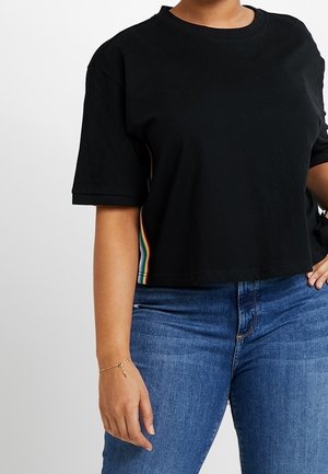 Camiseta negra estrecha de manga corta con rayas laterales arcoíris, usada con vaqueros azules, mostrada desde la mitad del cuello hasta las caderas, brazo izquierdo relajado.