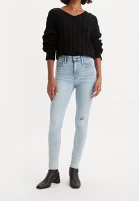 Suéter de punto negro con escote en V, combinado con jeans ajustados azul claro con un detalle desgastado, terminado con botas negras hasta el tobillo.