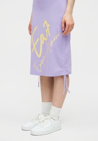 Κάτω μέρος σώματος ενός ατόμου που φοράει λιλά φούστα με κίτρινο κείμενο "Emporio Armani" και λευκά αθλητικά παπούτσια με λευκές καλτσες μπάσκετ.