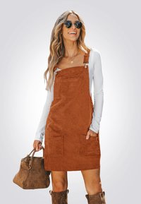 Rostfarbene Cordkleid mit verstellbaren Trägern und Fronttaschen, getragen über einem weißen Langarmoberteil; ergänzt durch eine braune Handtasche.