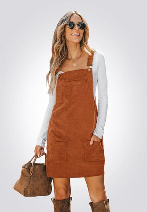 Rust-colored corduroy dress with adjustable straps, front pockets, worn over a white long-sleeve top; complemented by brown handbag.
