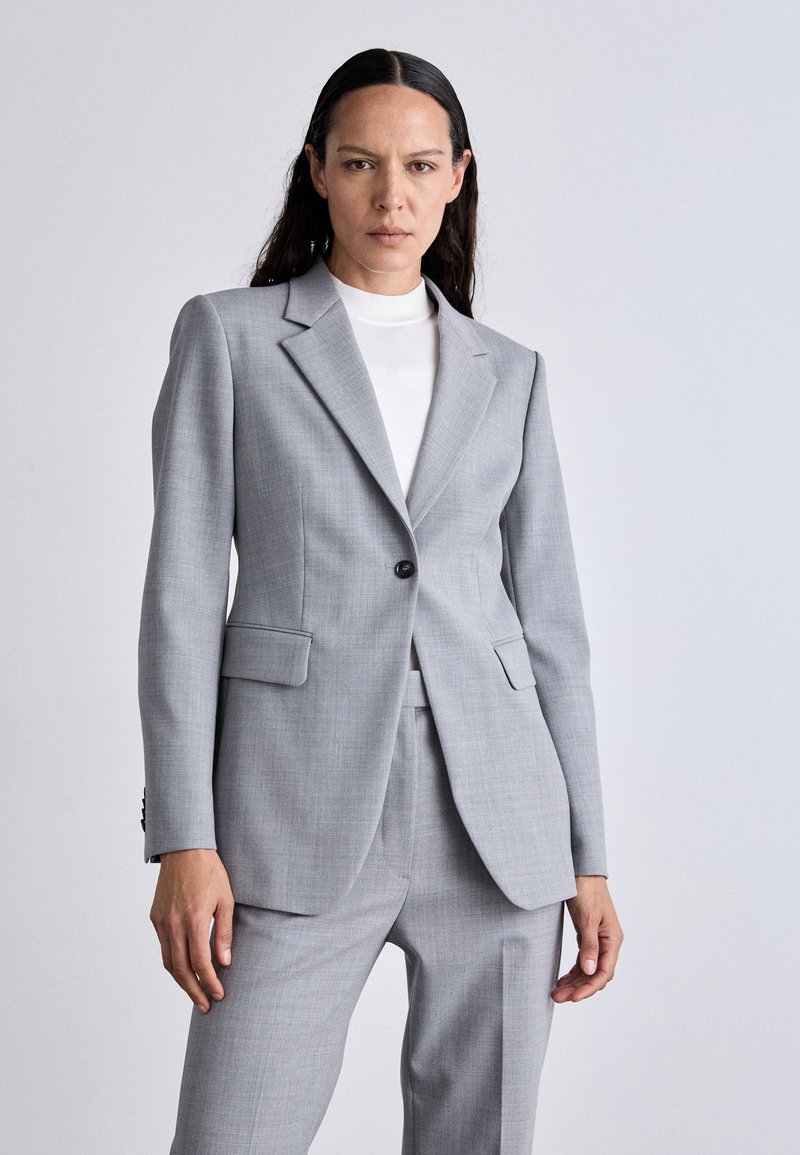 Fato cinza claro com um blazer estruturado, apresentando um botão simples, lapelas recortadas e dois bolsos frontais. Textura de tecido suave e ajuste sob medida.