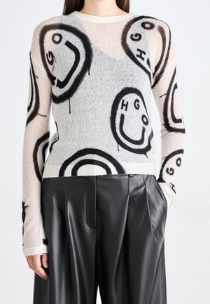 Pull beige clair avec des visages souriants abstraits noirs et des lettres, associé à un pantalon large en cuir noir taille haute.